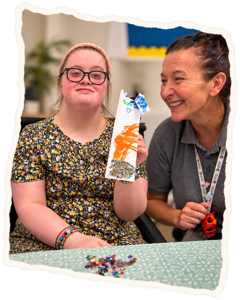 A young woman holding up a painted craft beside a smiling member of staff