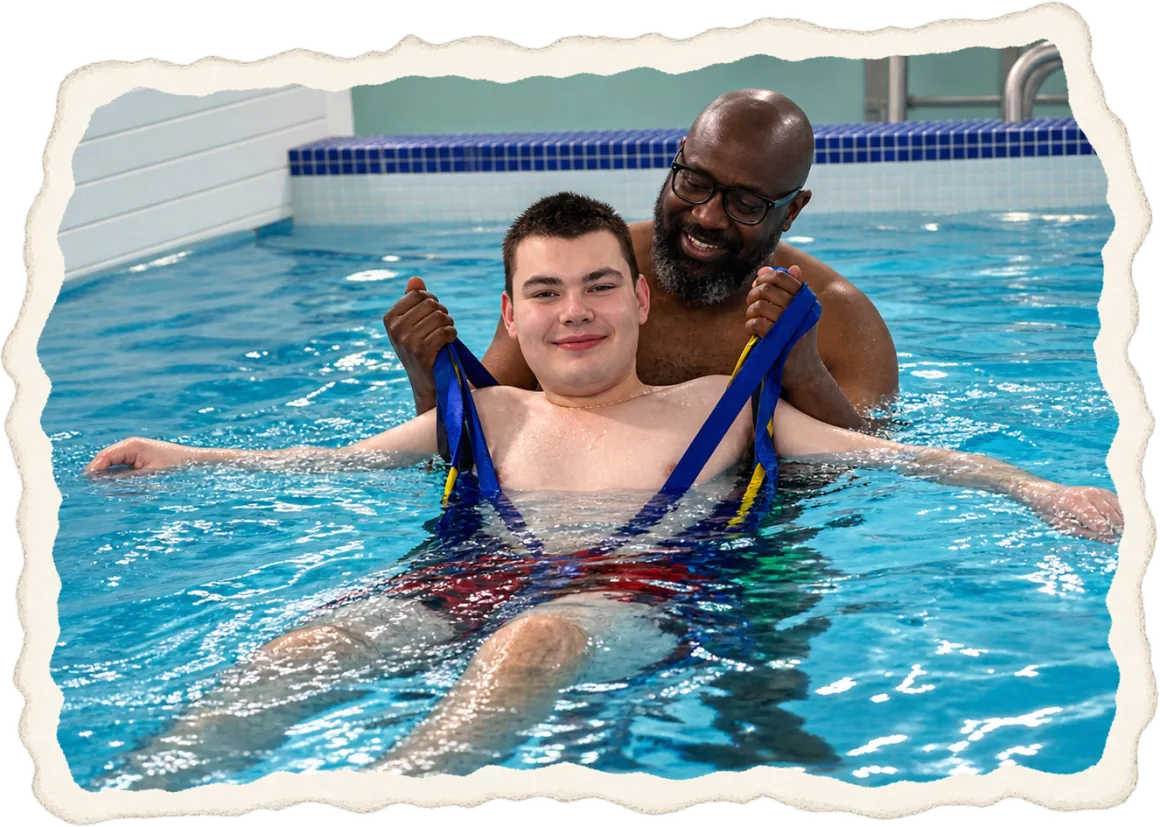 A young man floating in the hydropool in a blue support sling, steadied from behind by a smiling helper