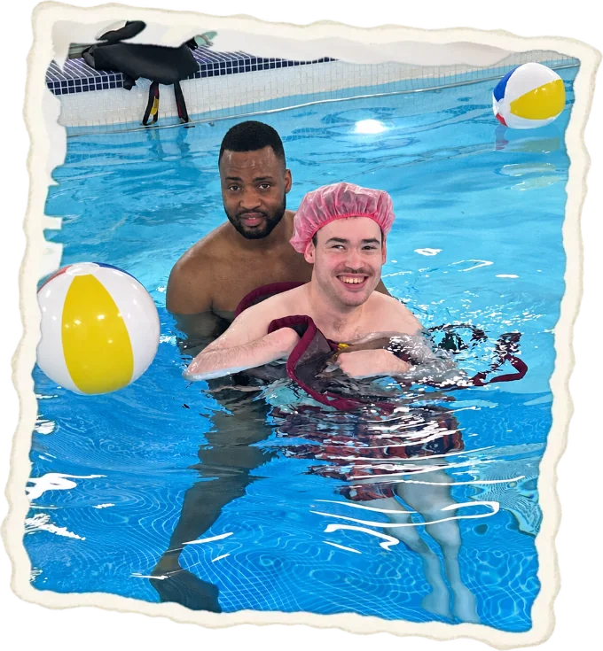A swimmer in a pink cap in the hydropool with a support worker, a beach ball floating beside them