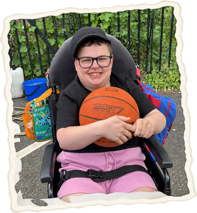 A young man in his wheelchair hugging a basketball, grinning at the camera